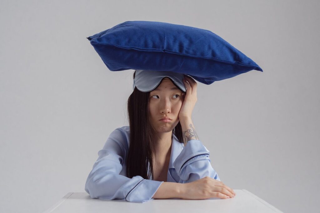 Mulher com expressão de insatisfação trajada de pijamas e com uma máscara de dormir posicionada na testa (acima dos olhos), com um travesseiro apoiado na cabeça