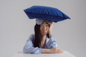 Mulher com expressão de insatisfação trajada de pijamas e com uma máscara de dormir posicionada na testa (acima dos olhos), com um travesseiro apoiado na cabeça