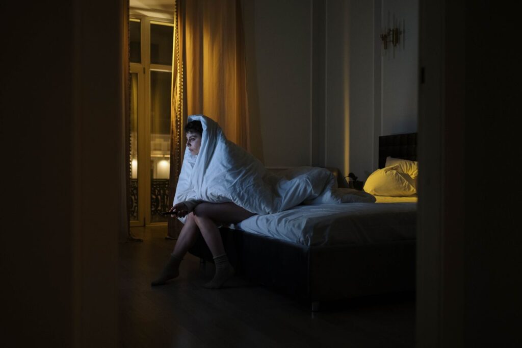Mulher sentada na beira da cama acordada e assistindo televisão, enrolada em um cobertor, sendo iluminada pela luz da televisão, com o controle remoto na mão